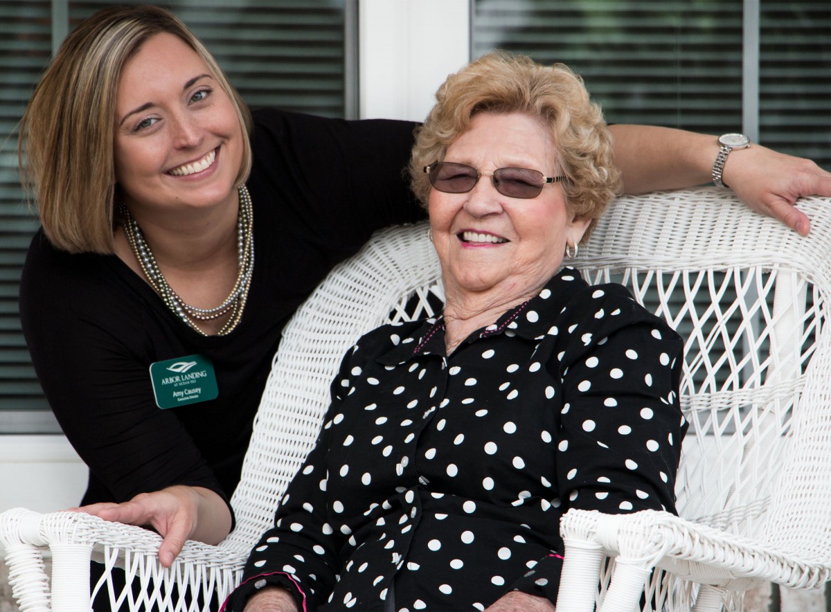 A smiling elderly woman in a polka dot shirt sits on a white wicker chair next to a younger woman outdoors.