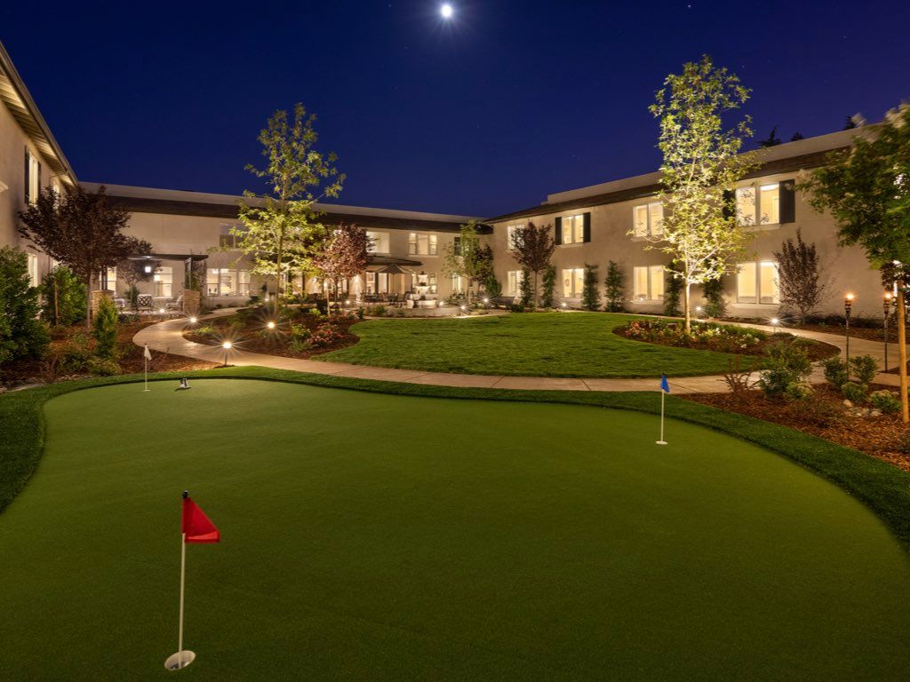 A serene night view of a well-lit putting green surrounded by a building and trees.