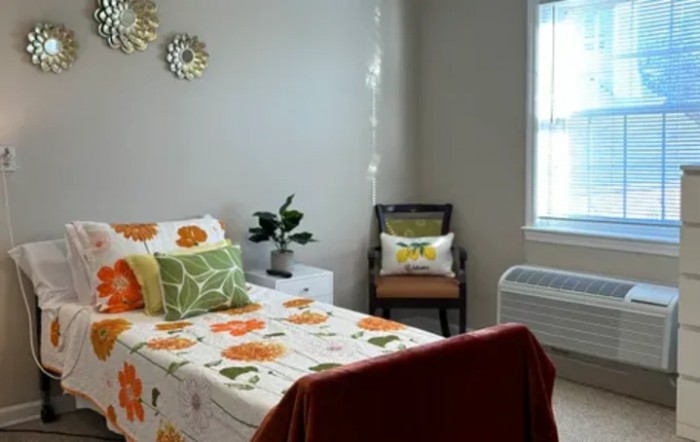 A cozy bedroom with a floral-themed single bed, featuring decorative pillows and a small chair by the window.