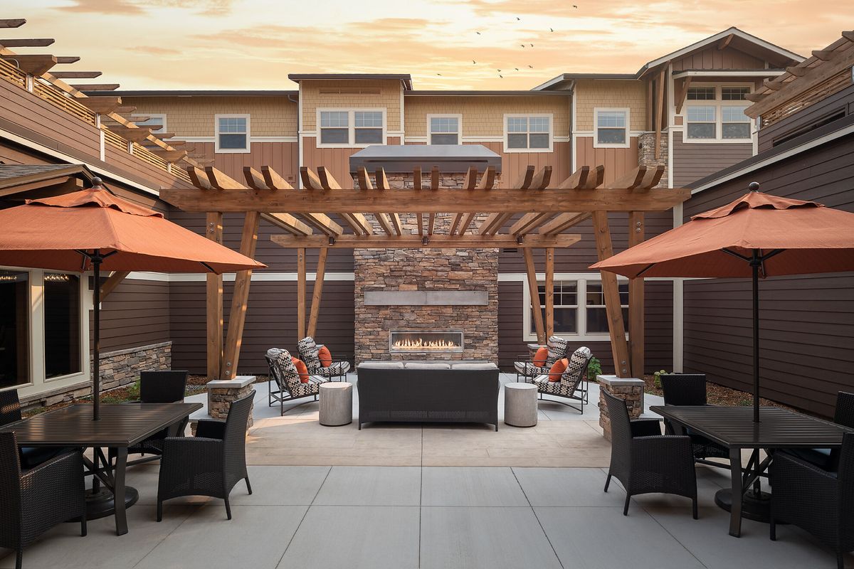 A modern outdoor patio with a pergola and central fireplace surrounded by seating and umbrellas.