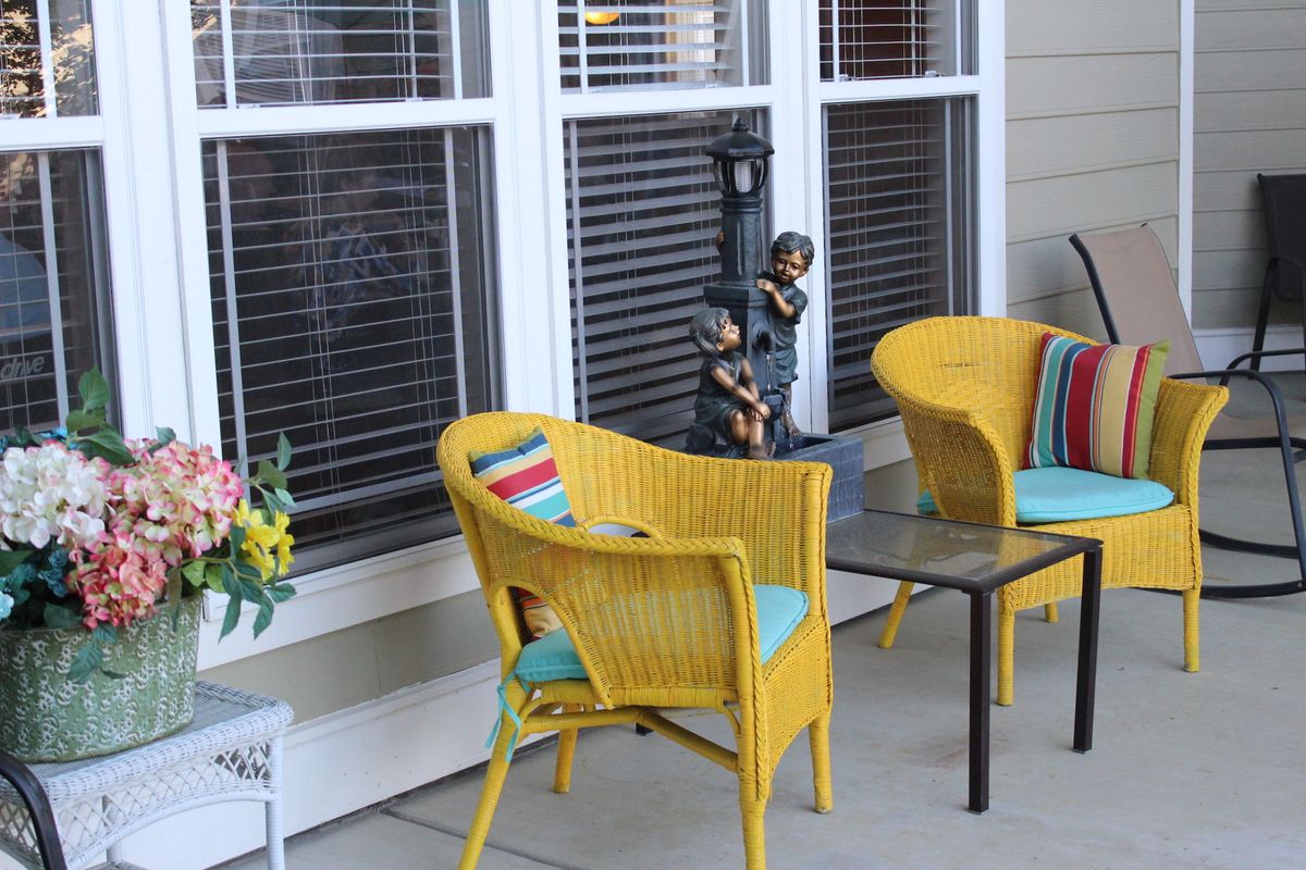 A cozy patio setup with yellow wicker chairs and colorful cushions, accompanied by a decorative flower arrangement beside a window.
