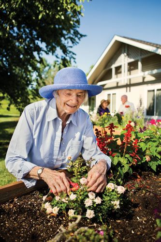 An elderly woman wearing a blue sun hat is happily tending to her garden in front of a house, surrounded by blooming flowers.
