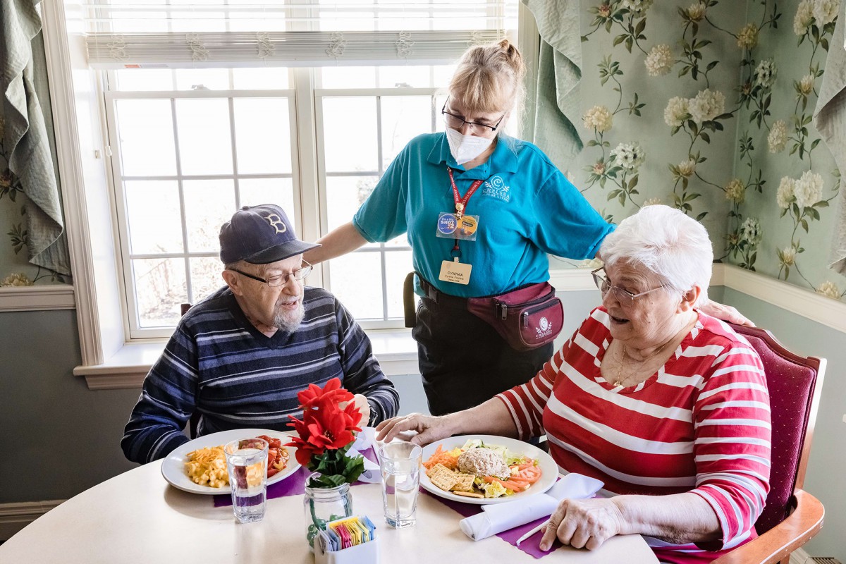A caregiver assists two elderly individuals during a meal in a nursing home setting.