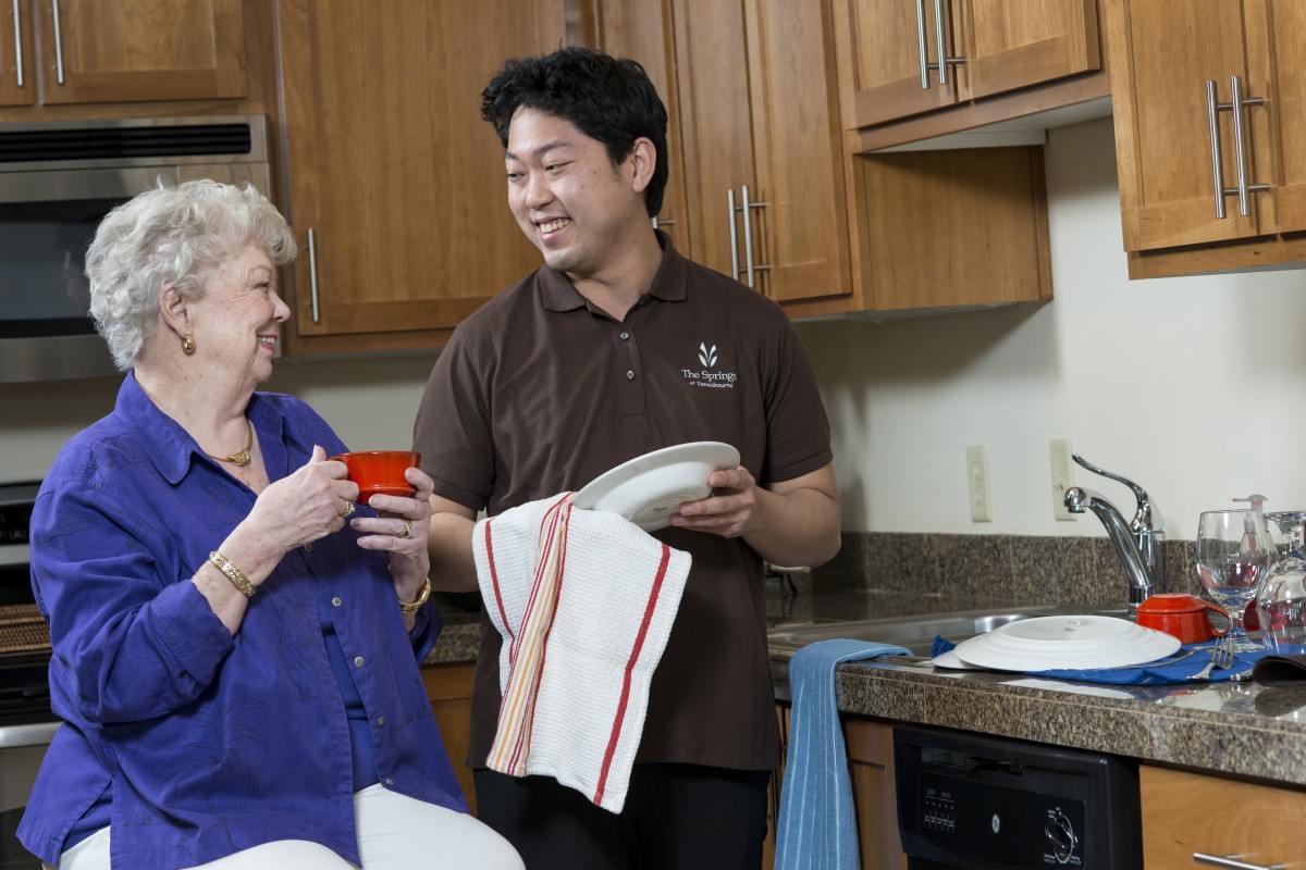 A person is helping an older adult with household chores in the kitchen.