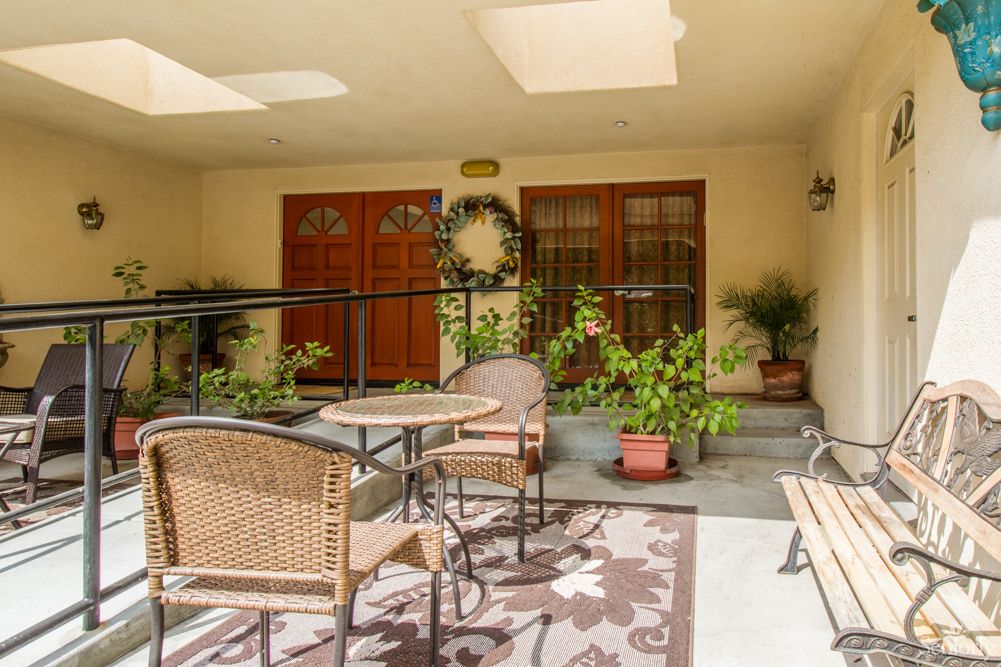 A cozy patio with wicker chairs and a table surrounded by potted plants, featuring wooden doors adorned with a decorative wreath under warm sunlight.