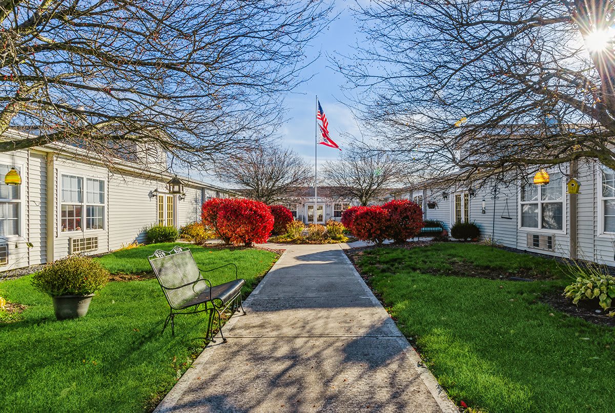 A scenic outdoor pathway leads to a building, flanked by benches and vibrant red bushes, with a flag waving in the background.