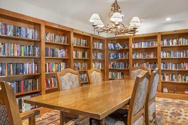 A cozy library room with wooden shelves filled with books and a large table surrounded by chairs.