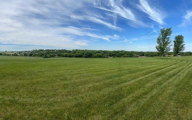 A vast green field with a clear blue sky and scattered clouds.