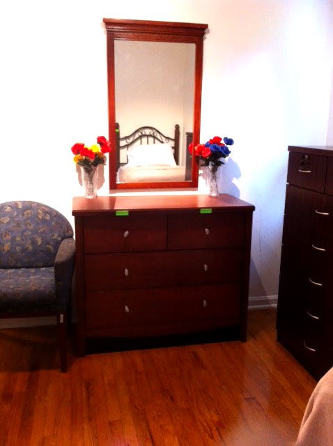 A room interior featuring a wooden dresser with a mirror, flanked by vases of colorful flowers, next to a cushioned chair on a wooden floor.
