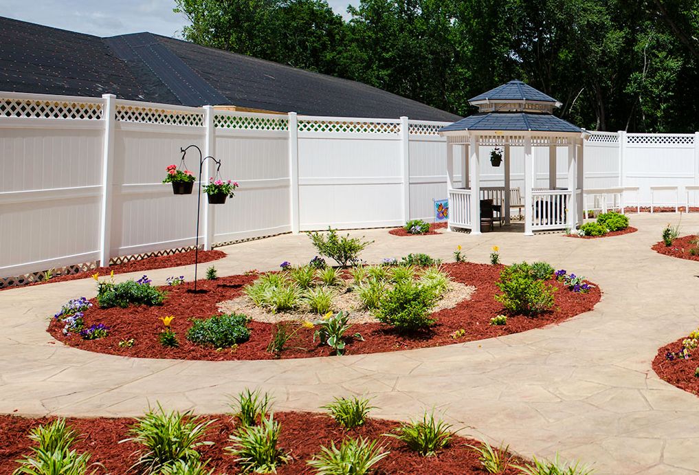 A beautifully landscaped garden with a central gazebo, surrounded by mulched flower beds and a white fence.
