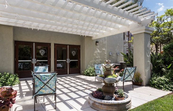 A cozy outdoor patio featuring a pergola casting shadows, with a central fountain surrounded by chairs.