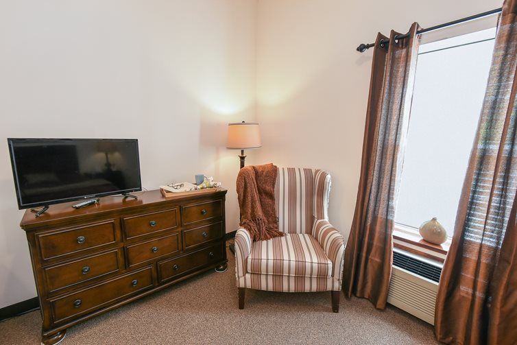 A cozy living room corner featuring a striped armchair, a wooden dresser with a TV, and soft lighting from a floor lamp.