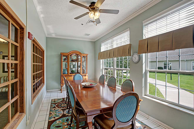 A well-lit dining room featuring a long wooden table with matching chairs, large windows with curtains, and a decorative cabinet.