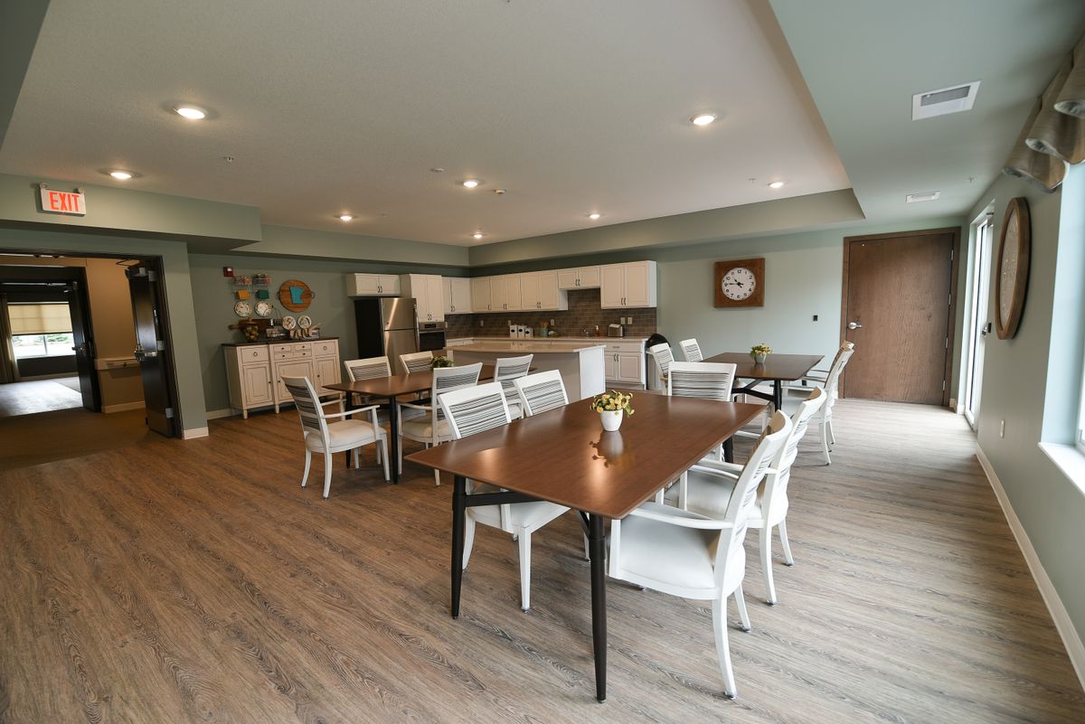 A spacious dining area with multiple tables and chairs, featuring a modern kitchen in the background and wood flooring.