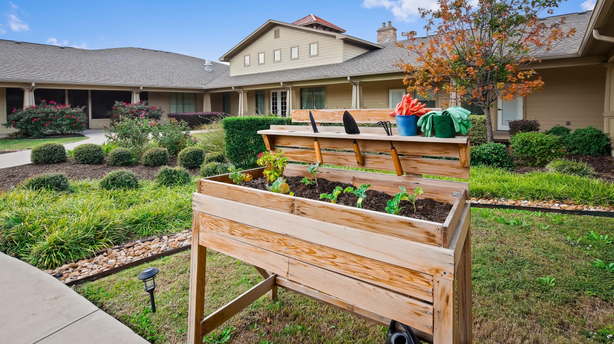 A backyard garden with a raised wooden planter box and gardening tools in front of a house.