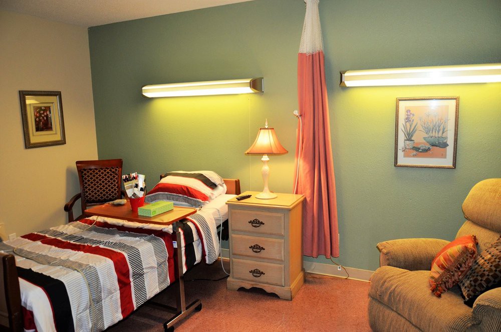 A hospital-style bedroom with a single bed, nightstand, lamp, and chair.