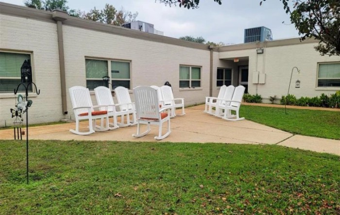 A patio area with white chairs arranged outside a building on a grassy lawn.