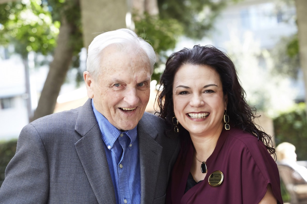 A smiling elderly man and a woman posing together outdoors.