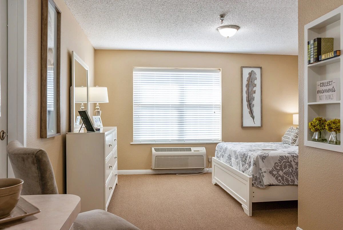 A cozy bedroom with neutral tones, featuring a bed, dresser, and window with blinds.
