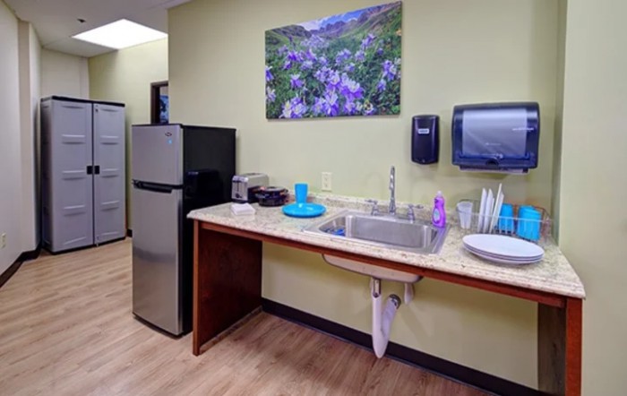 A small office kitchen area with a sink, countertop, refrigerator, and wall art.
