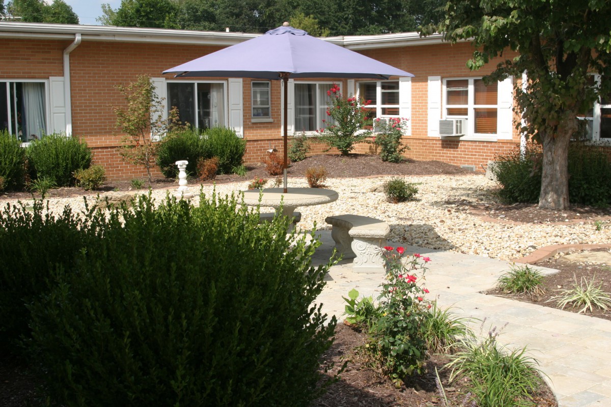 A peaceful garden patio with a large umbrella providing shade over a picnic table, surrounded by plants and a stone pathway.