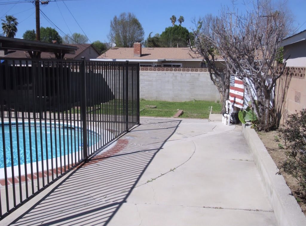 A backyard with a fenced swimming pool and patio area under a clear blue sky.