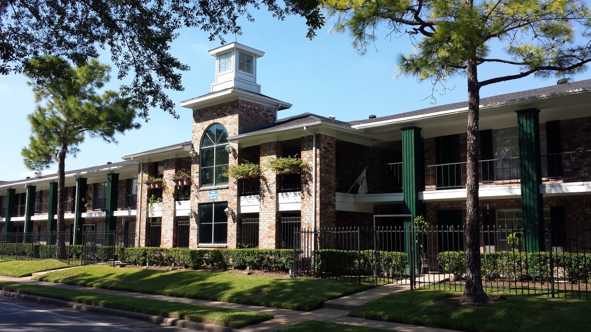 A two-story residential apartment building with a distinctive arched window in the center and surrounded by trees and a gated entrance.