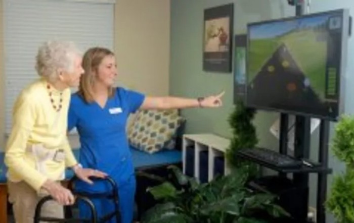 A healthcare professional assists an elderly patient with physical therapy using a video screen.