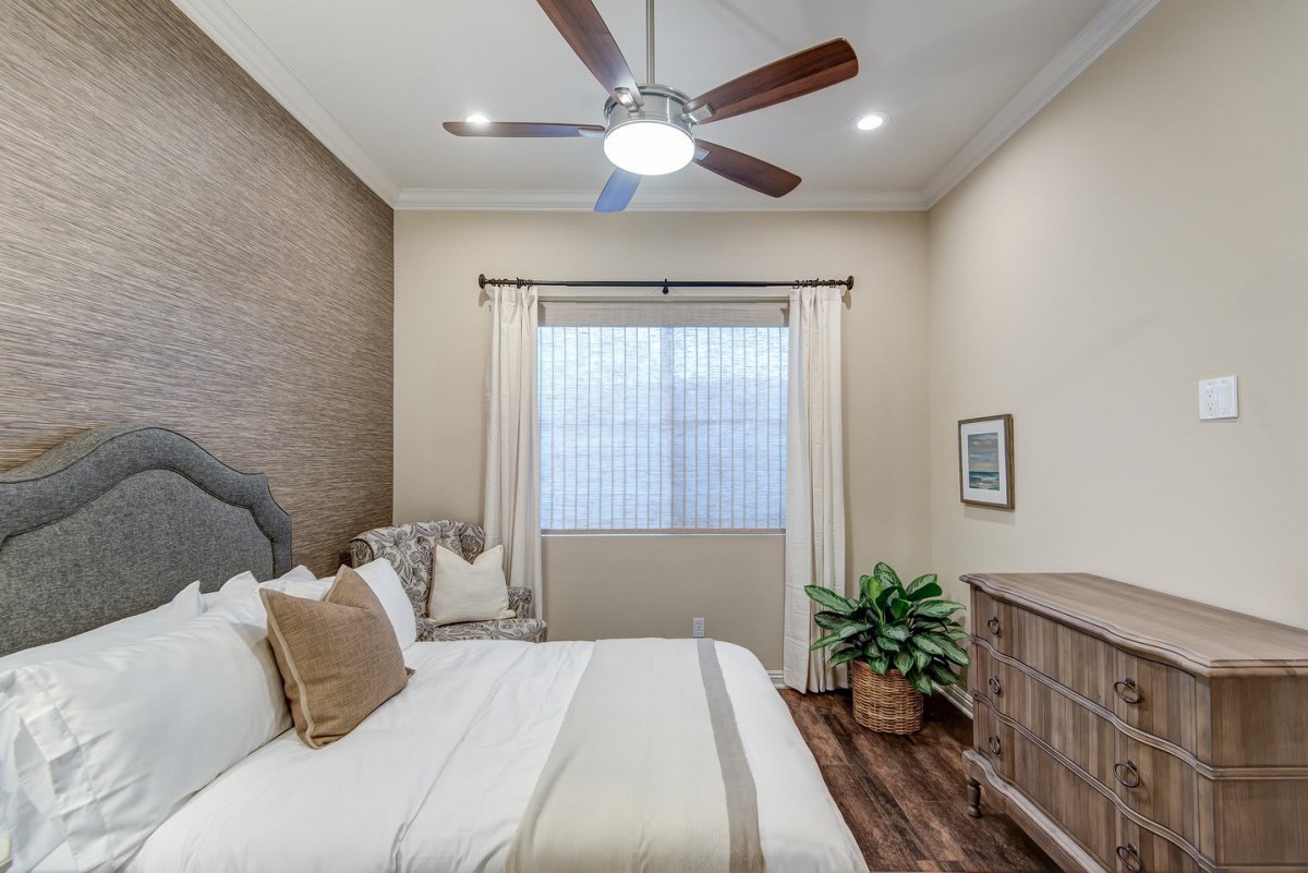 A modern bedroom with neutral tones, featuring a ceiling fan, bed, window with curtains, a plant, and a dresser.