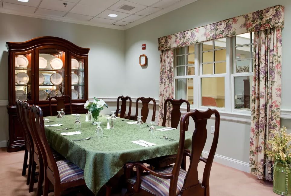 An elegant dining room with a long table covered by a green tablecloth, surrounded by wooden chairs, and a china cabinet in the background.