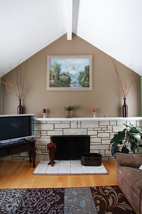 A cozy living room with a central fireplace, adorned with a landscape painting and flanked by decorative vases. A television sits to the side, and an indoor plant adds a touch of greenery.