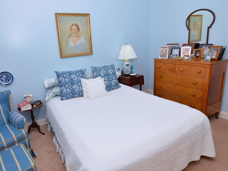 A cozy bedroom with blue walls, featuring a neatly made bed adorned with decorative pillows, a bedside lamp, a dresser with a mirror, and a portrait on the wall.