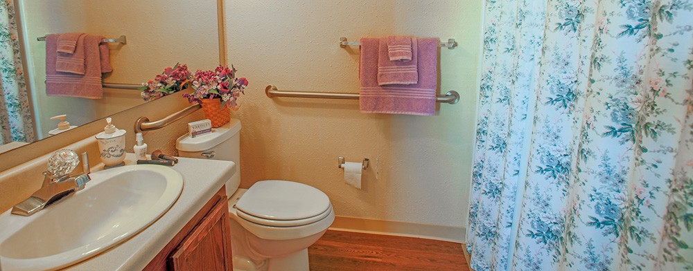 A cozy bathroom with a toilet, pink towels, grab bars, and floral decorations.