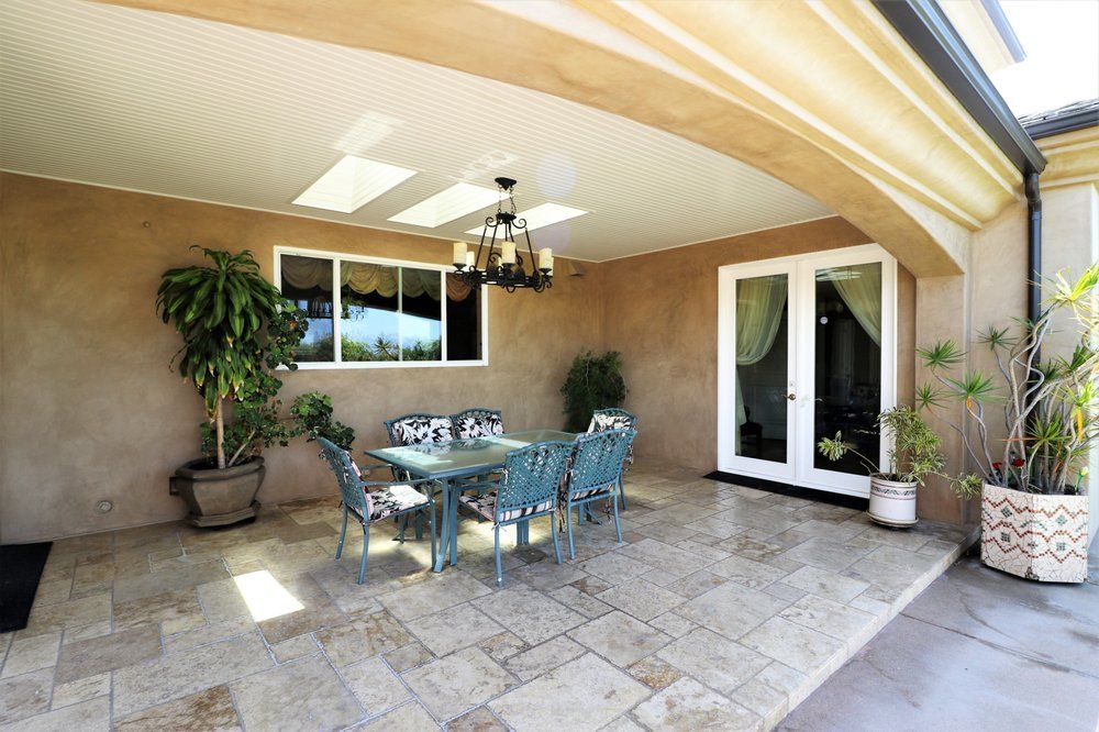 A spacious outdoor patio featuring a dining area with a chandelier, surrounded by potted plants and natural light.