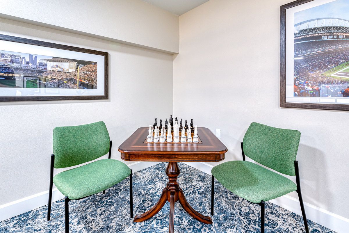A chess board set up on a small wooden table with two green chairs, surrounded by framed artwork.