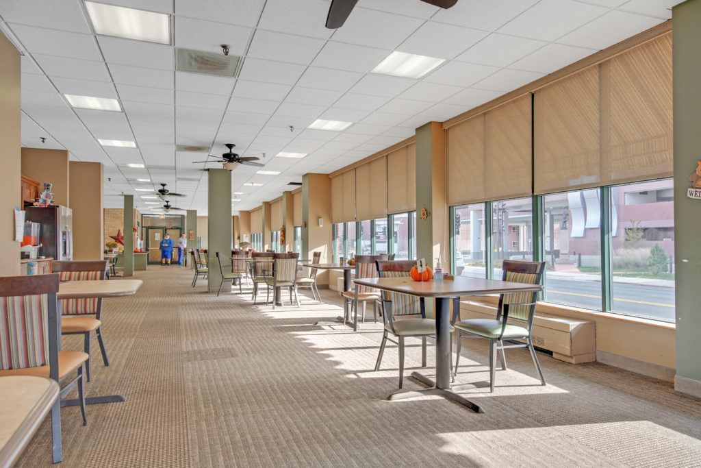 A spacious dining area with tables and chairs, featuring large windows with blinds and ceiling fans.
