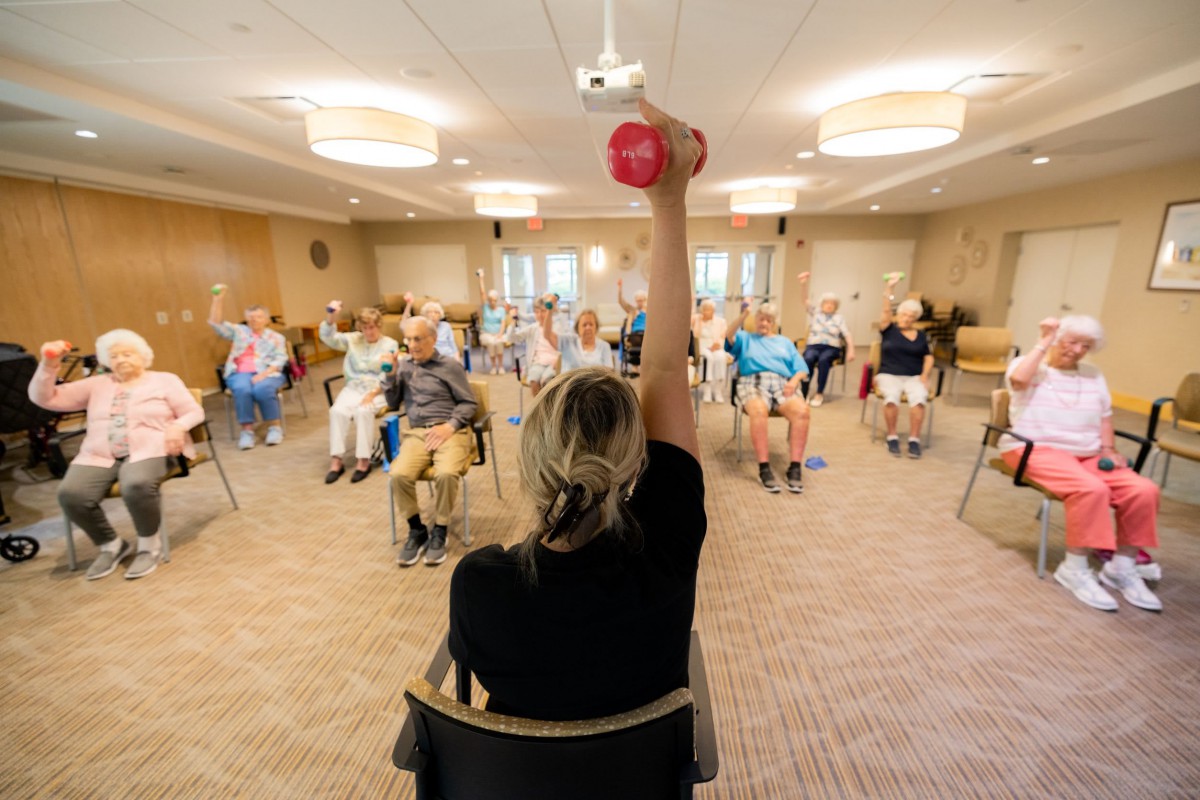 A group of seniors participating in a seated exercise class led by an instructor holding a dumbbell.