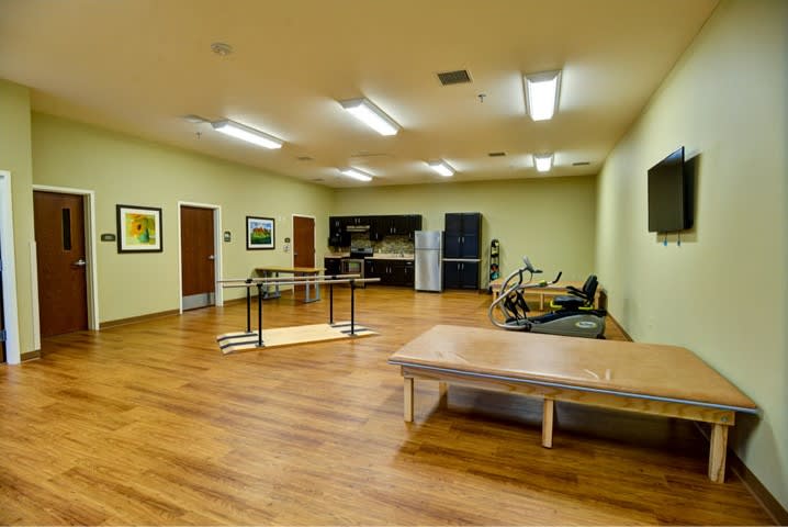 A rehabilitation room equipped with exercise machines and therapy tables on a wooden floor.