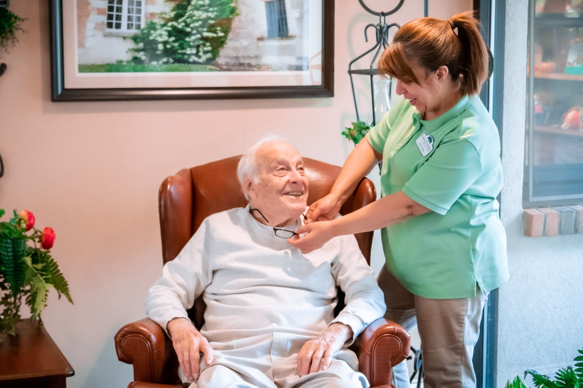 A caregiver assisting an elderly man seated in a comfortable chair.