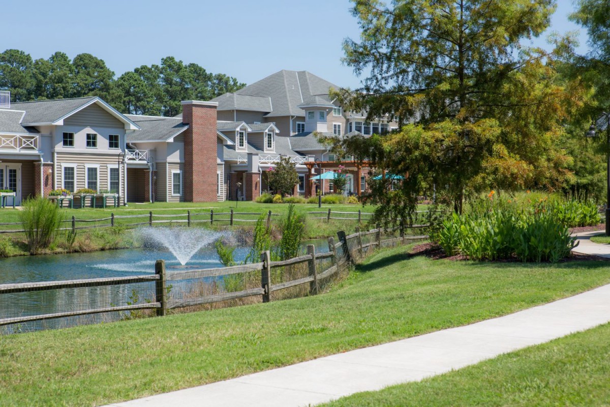 A scenic residential area featuring a pond with a fountain, surrounded by greenery and houses.