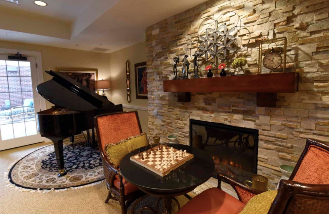 A cozy living room with a chess set on a table, featuring a fireplace with a stone wall and a grand piano in the background.