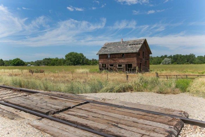 An abandoned wooden house stands in a rural landscape under a clear blue sky.
