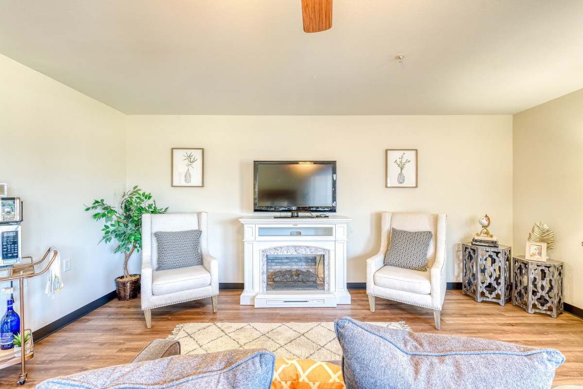 A cozy living room with a television above a white fireplace, flanked by two armchairs and decorated with plants and artwork.