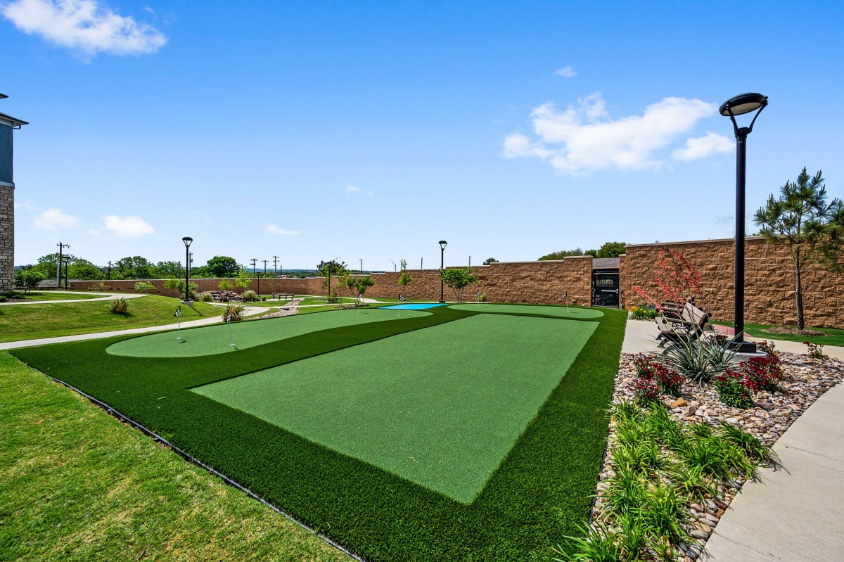 A landscaped outdoor area with artificial turf, surrounded by greenery and a clear blue sky.
