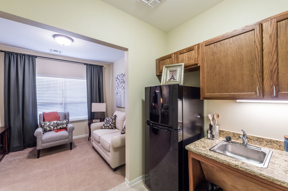 A cozy apartment featuring a compact kitchen with wooden cabinets and a black fridge, opening into a comfortable living room with a couch and armchair.