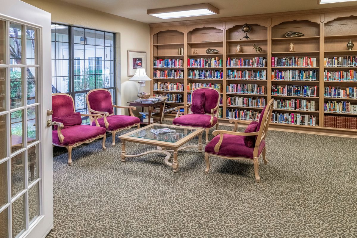 A cozy library reading room with bookshelves, plush seating, and a central table.