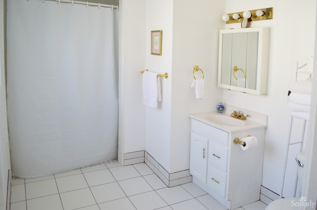 A bright bathroom with white tiles, a vanity with a mirror, and gold-accented fixtures.