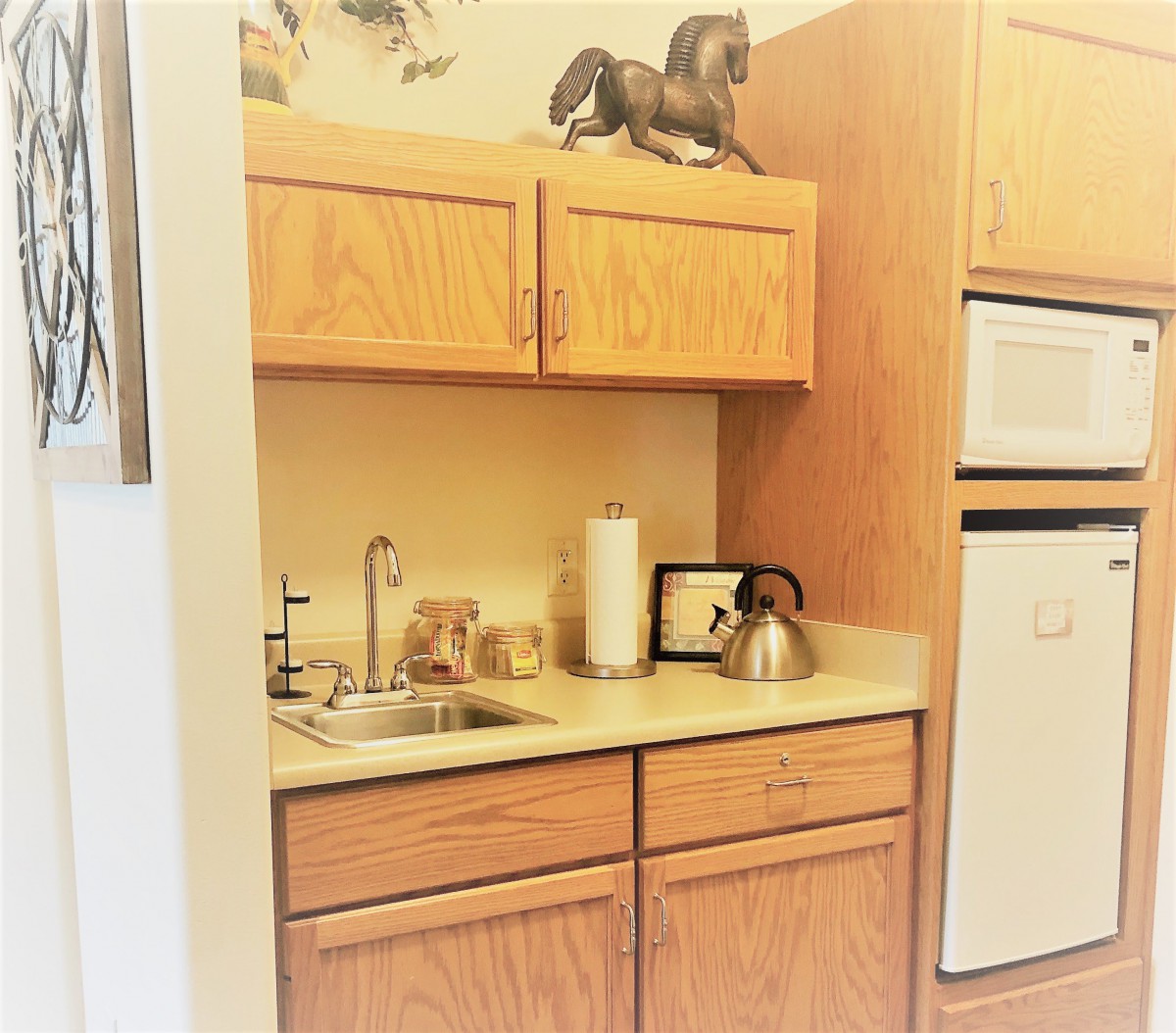 A small kitchen area with wooden cabinets, a countertop featuring a sink and kettle, and a decorative horse on the upper cabinet.