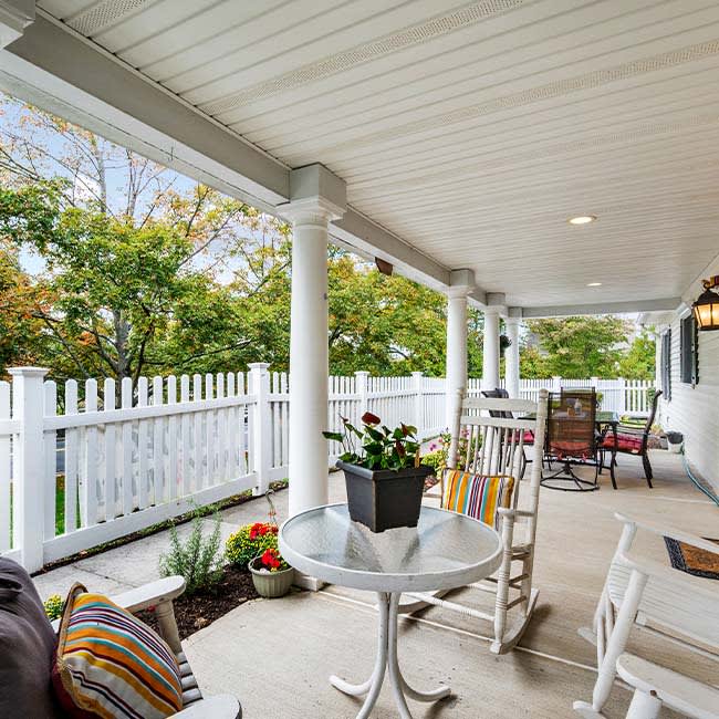 A cozy porch area with rocking chairs, a table, and plants, all under a covered outdoor space with a white picket fence surrounding it.