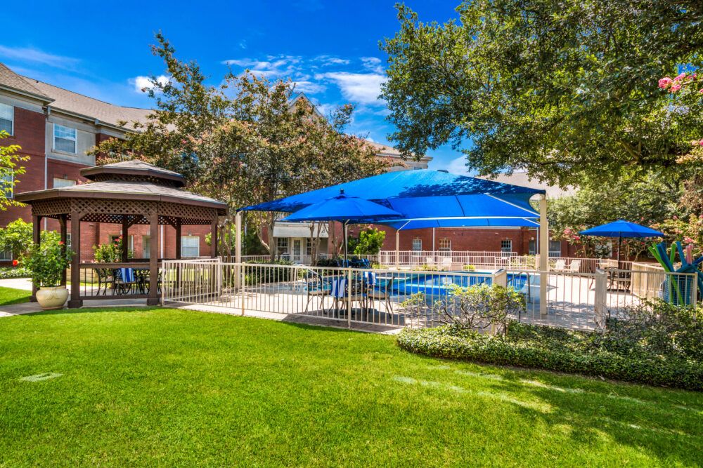 A well-maintained outdoor area featuring a pool with a blue canopy, surrounded by lush greenery and a gazebo under a clear blue sky.